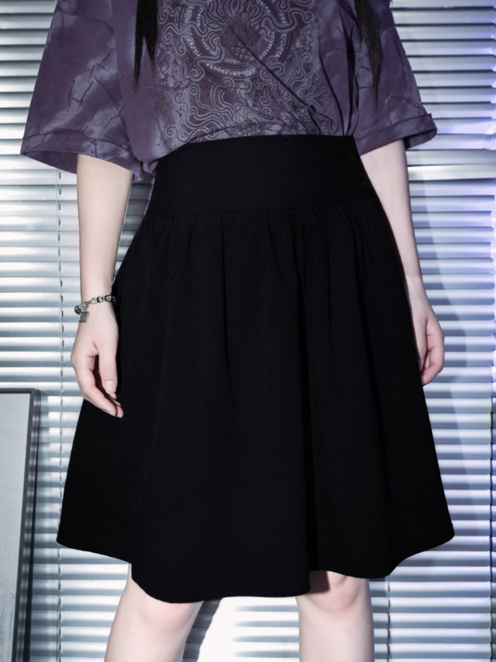 A full-length view of a model showcasing a black dark academia midi skirt with a thick high waistband and gathered pleats, styled with a purple cyber grunge top and a silver metallic hardware belt.