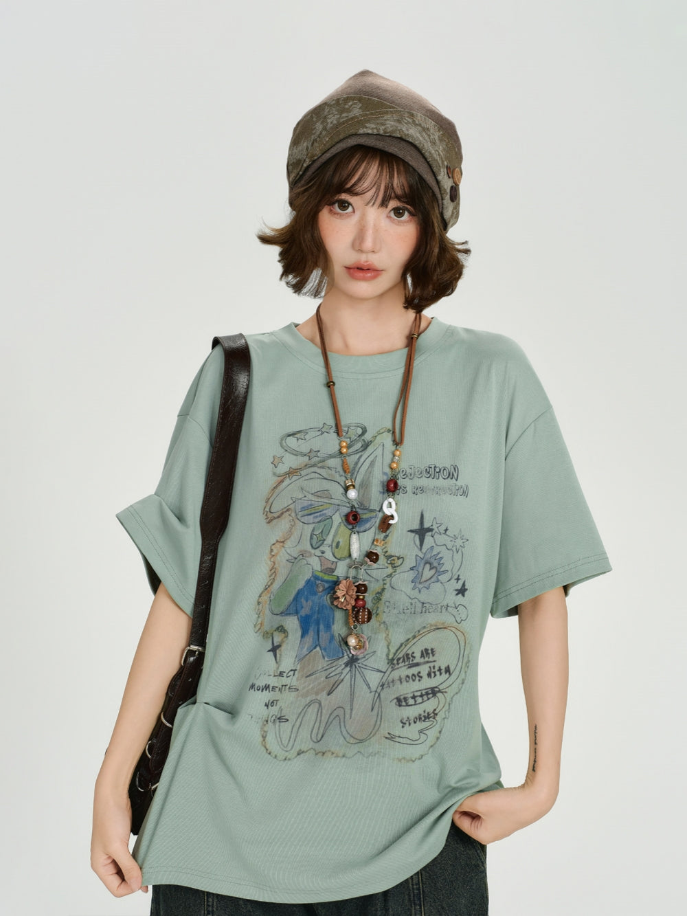 Oversized sage green t-shirt featuring a vintage hand-drawn cartoon graphic with whimsical doodles and artistic text.