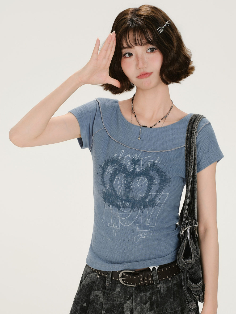 A close-up view of the same blue fairy-grunge distressed baby tee, showing the detail of the raw-edge seams, the ribbed texture, and the grunge crown graphic. The model holds one hand to her forehead in a nonchalant gesture. She is accessorized with layered necklaces. The image clearly displays the top's fit and worn-in appearance.