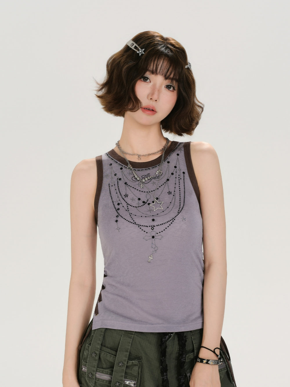 A close-up shot of a model wearing a fairy-grunge side lace-up tank in distressed purple. The tank features brown ribbed binding, a dark graphic of layered chains with beaded cross and star accents. The side view shows the brown cord lace-up detail. She wears it with layered necklaces and green cargo pants.