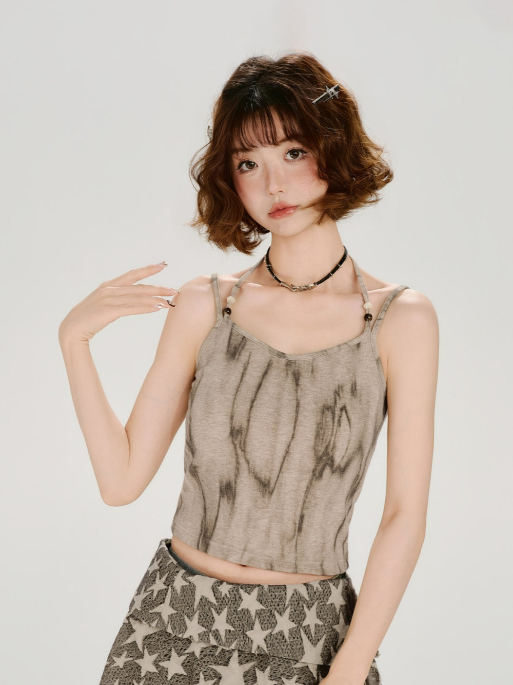 Front view of a model wearing a fairy-grunge distressed halter tank. The top features a vertical earth-toned tie-dye pattern on a sand-colored base, with thin halter straps detailed with small beads. She is styled with a layered star-print skirt and a dark choker.