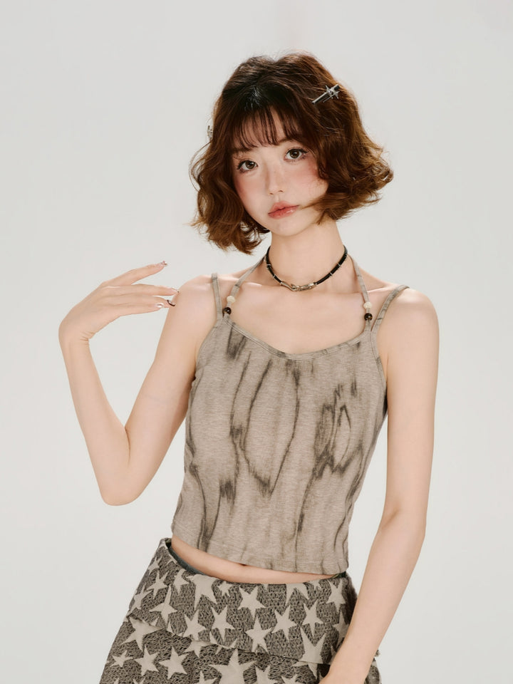 Front view of a model wearing a fairy-grunge distressed halter tank. The top features a vertical earth-toned tie-dye pattern on a sand-colored base, with thin halter straps detailed with small beads. She is styled with a layered star-print skirt and a dark choker.