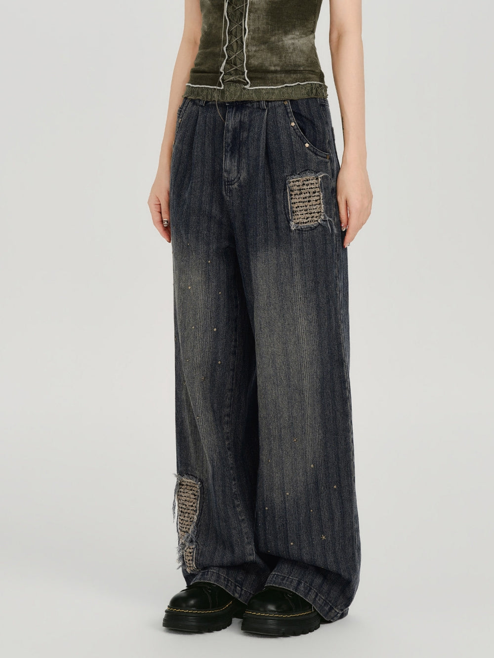 A product showcase of 90s grunge distressed patchwork jeans featuring a deep indigo wash with vertical fading. The jeans are detailed with frayed woven tapestry patches on the pocket and lower leg, accented by subtle metallic studs. The model pairs them with a lace-up olive corset top and chunky black boots for a complete wasteland aesthetic.
