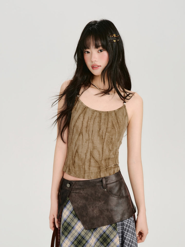 A product showcase of a fairy-grunge acid wash camisole in khaki and dusty pink tones. The top features a unique crinkled, vertical ruched texture and thin spaghetti straps with bead accents. It is styled in a 90s grunge aesthetic with low-rise distressed denim, a pleated leather skirt, and layered necklaces, emphasizing a worn-in, rebel vibe.