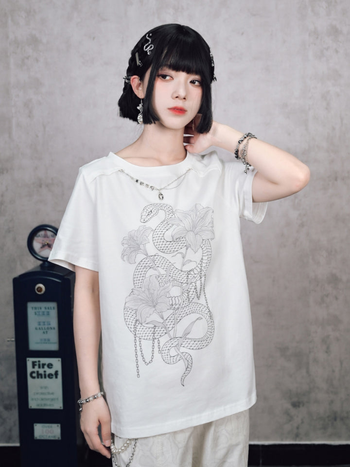 A model wearing a cyber grunge aesthetic graphic tee in white, featuring a serpent and floral print with a built-in neck chain, posed against industrial metallic blinds.