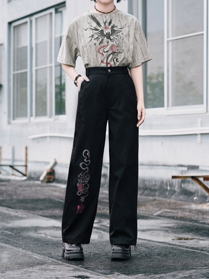 A full-length view of a model styling industrial grunge wide leg pants in black, featuring a detailed red and grey occult graphic print along the lower right leg, paired with platform punk boots.