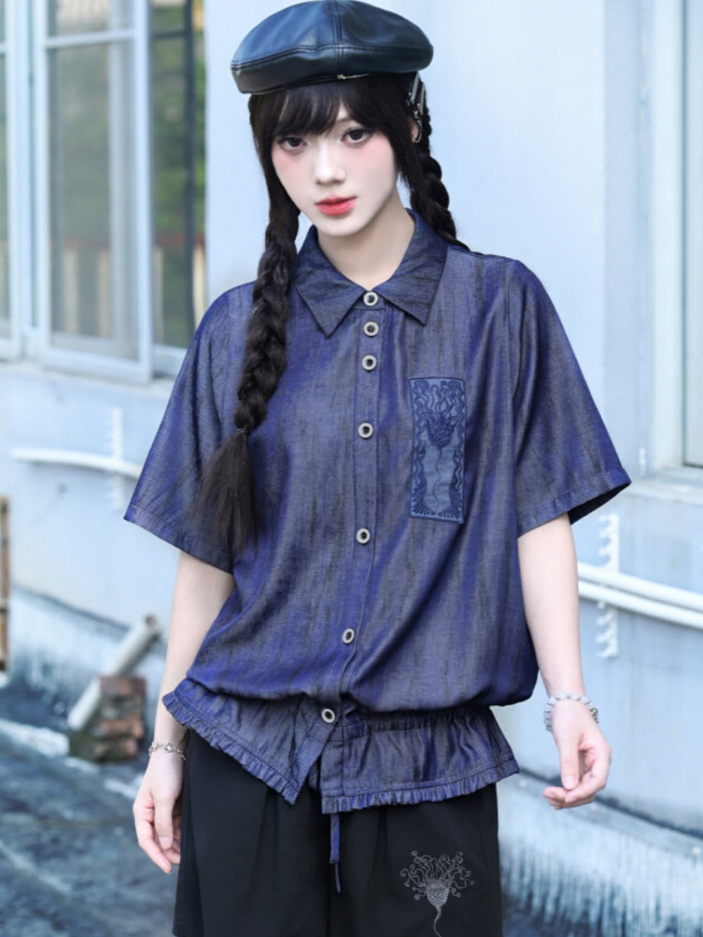A model wearing a metallic-wash blue cyber grunge button up shirt featuring a raw gathered elastic hem and a rectangular occult graphic patch on the left chest.