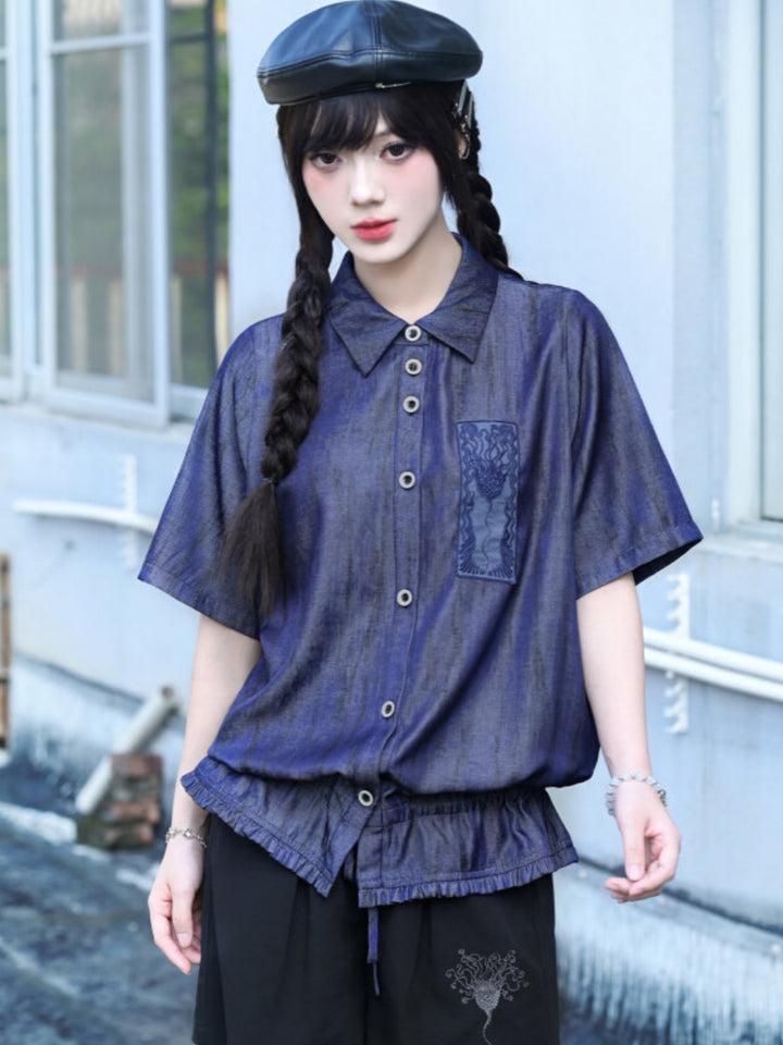 A model wearing a metallic-wash blue cyber grunge button up shirt featuring a raw gathered elastic hem and a rectangular occult graphic patch on the left chest.