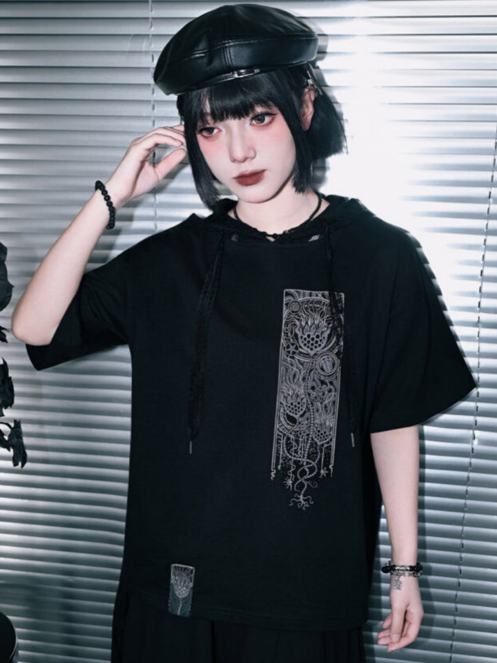 A full-length studio shot of a model styling a black industrial goth graphic tee featuring a built-in lace hood, drawstring neck, and a vertical mechanical occult print, paired with a layered maxi skirt.